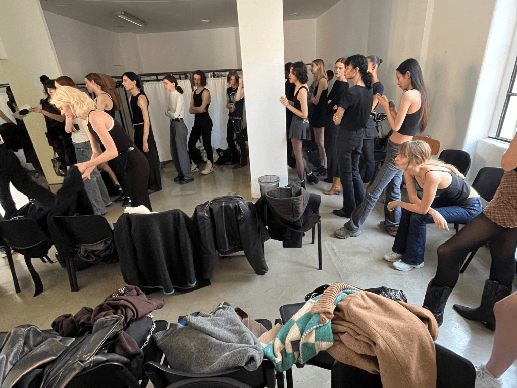 A group of models lined up in a dressing area, wearing black attire and waiting near a mirror. Clothing items are scattered on chairs in the foreground.