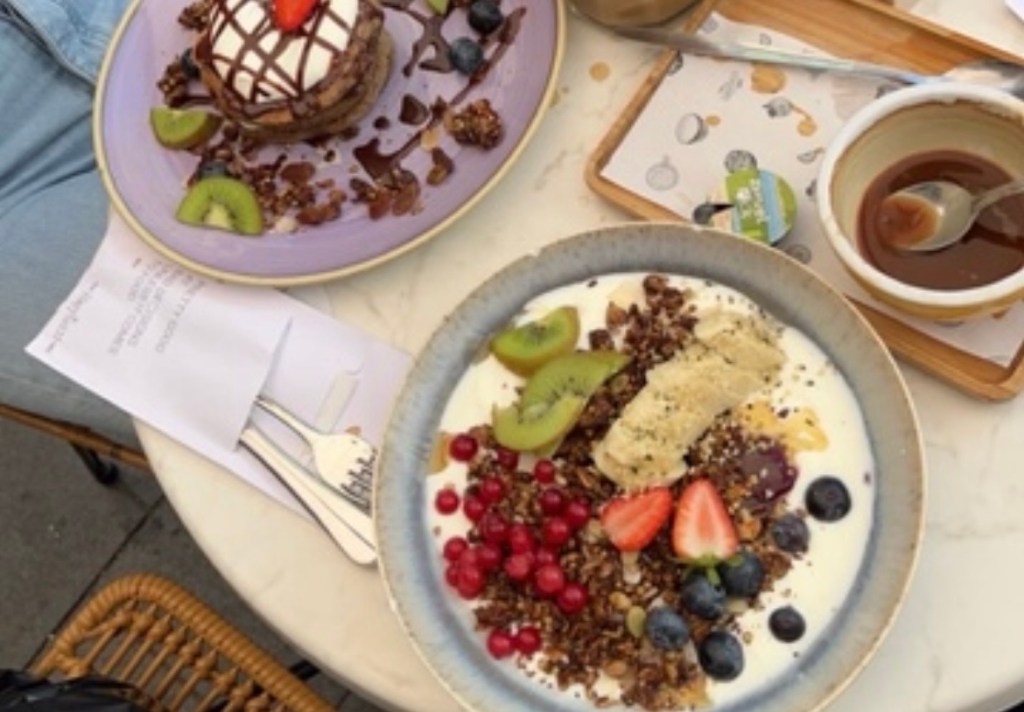 A table setting featuring two dessert plates. One plate has pancakes topped with whipped cream, chocolate drizzle, and strawberries, while the other features yogurt accompanied by granola, berries, and slices of kiwi and banana. A small dish of chocolate sauce is also visible on the table.